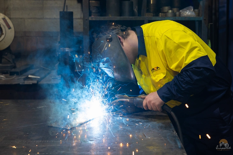 Welder in PPE welding parts on top of a metal bench.